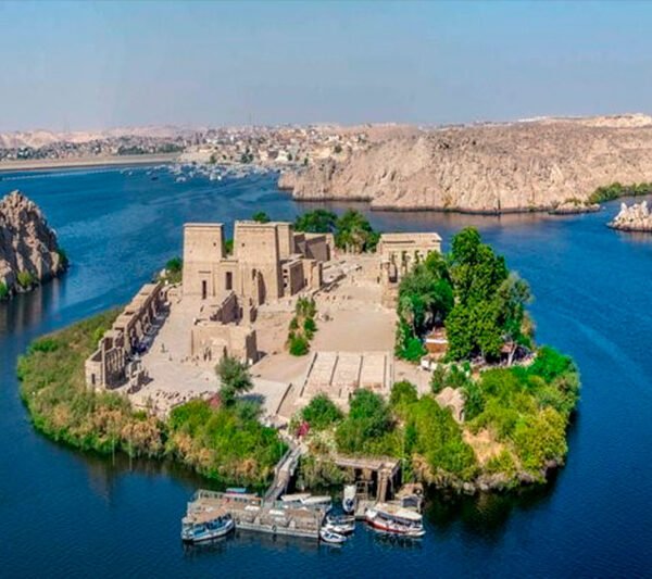 Philae Temple on a green island surrounded by blue Nile waters, with passenger boats at the landing