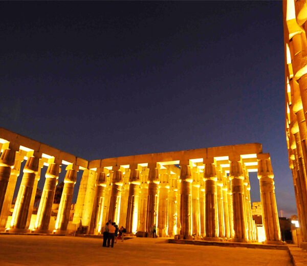 Visitors standing in a temple courtyard surrounded by rows of columns illuminated in golden light at night