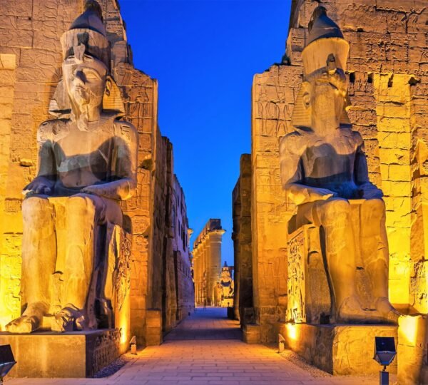 Two seated statues illuminated in gold at the entrance to Luxor Temple against a deep blue evening sky