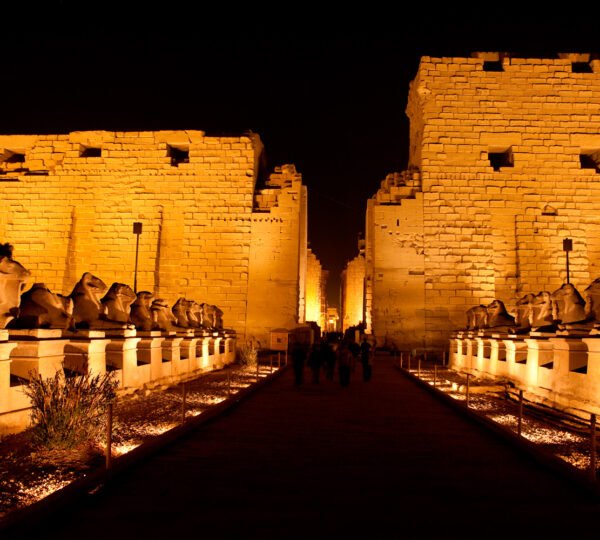 Rows of ram-headed sphinxes leading toward Karnak Temple’s golden illuminated entrance at night