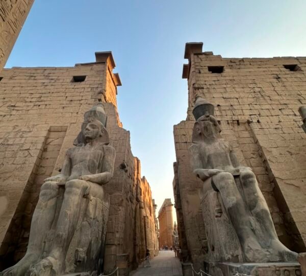 Two seated statues flanking the gateway between the pylons of Luxor Temple beneath a blue evening sky