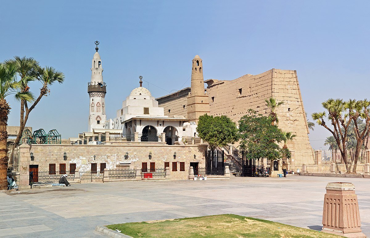 Abu Haggag Mosque and its minarets beside the ancient stone walls of Luxor Temple, with palm trees around the square