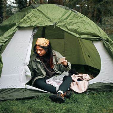 A camper seated at the entrance of a green tent in a wooded campsite