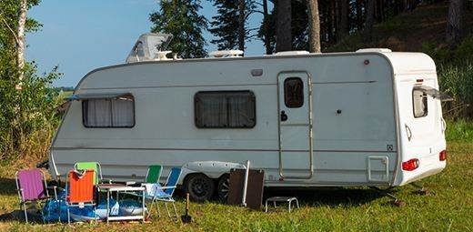 A white camper trailer parked beside trees with colorful camping chairs outside