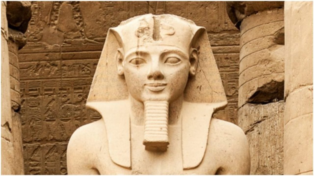 A pale stone royal statue with a carved headdress and long ceremonial beard in front of hieroglyph-covered masonry