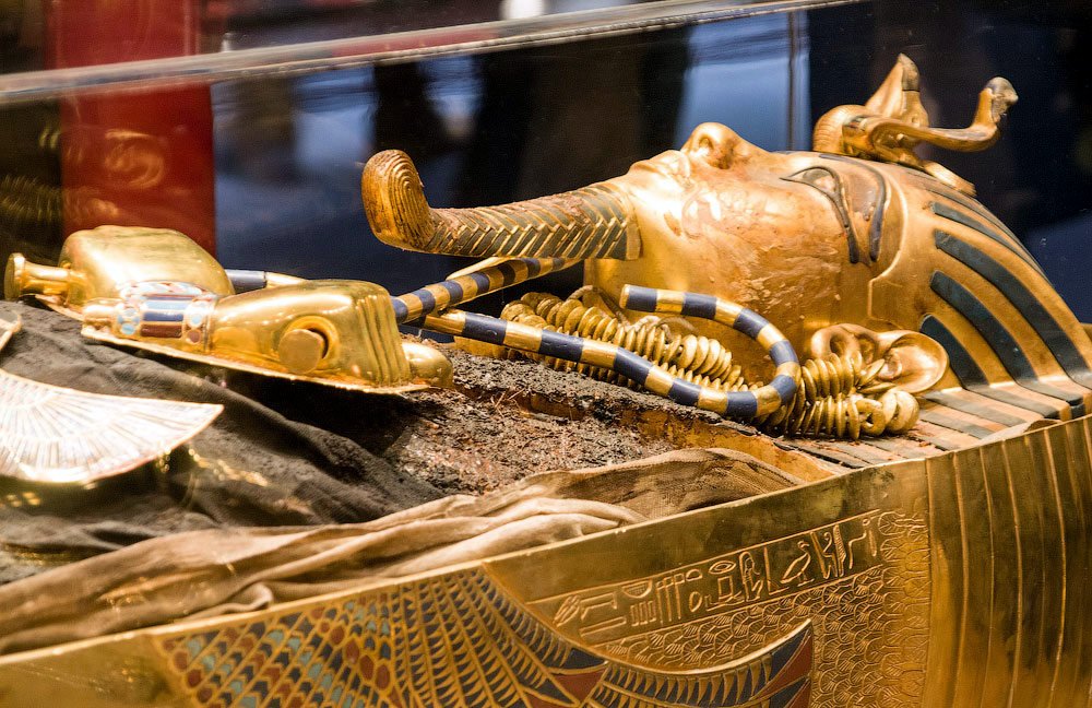 A golden royal funerary mask with a striped headdress, ceremonial beard and crossed crook and flail in a museum display