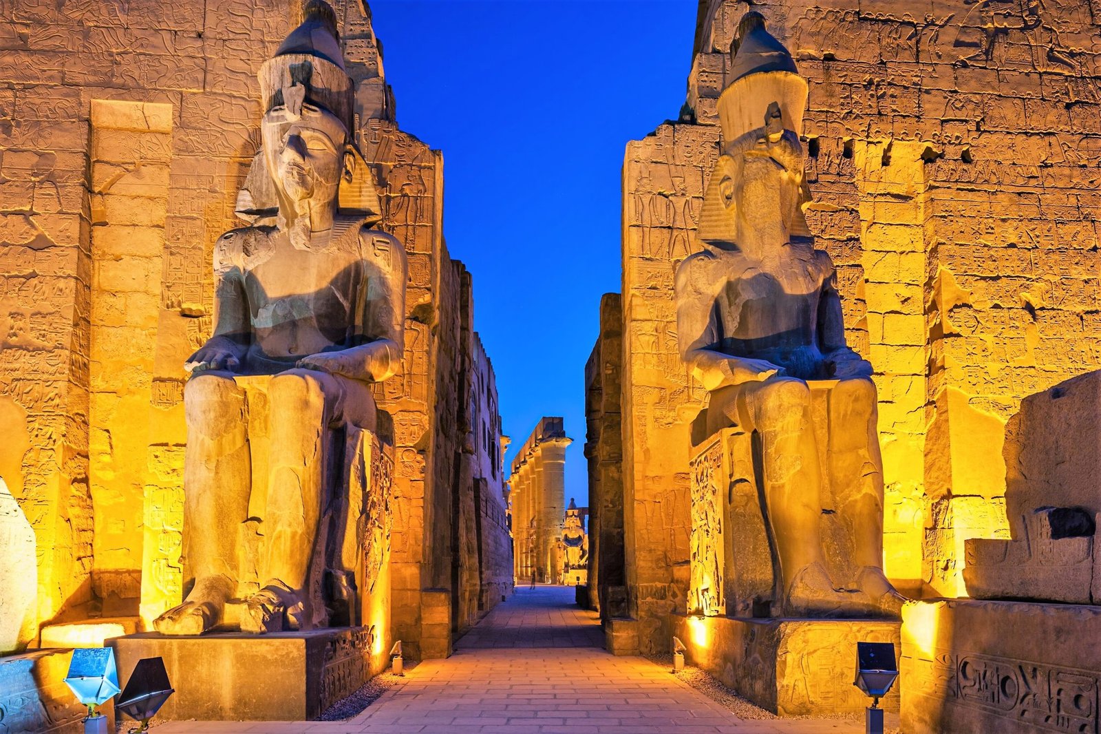 Two monumental seated statues illuminated in gold beside the entrance passage at Luxor Temple in the evening