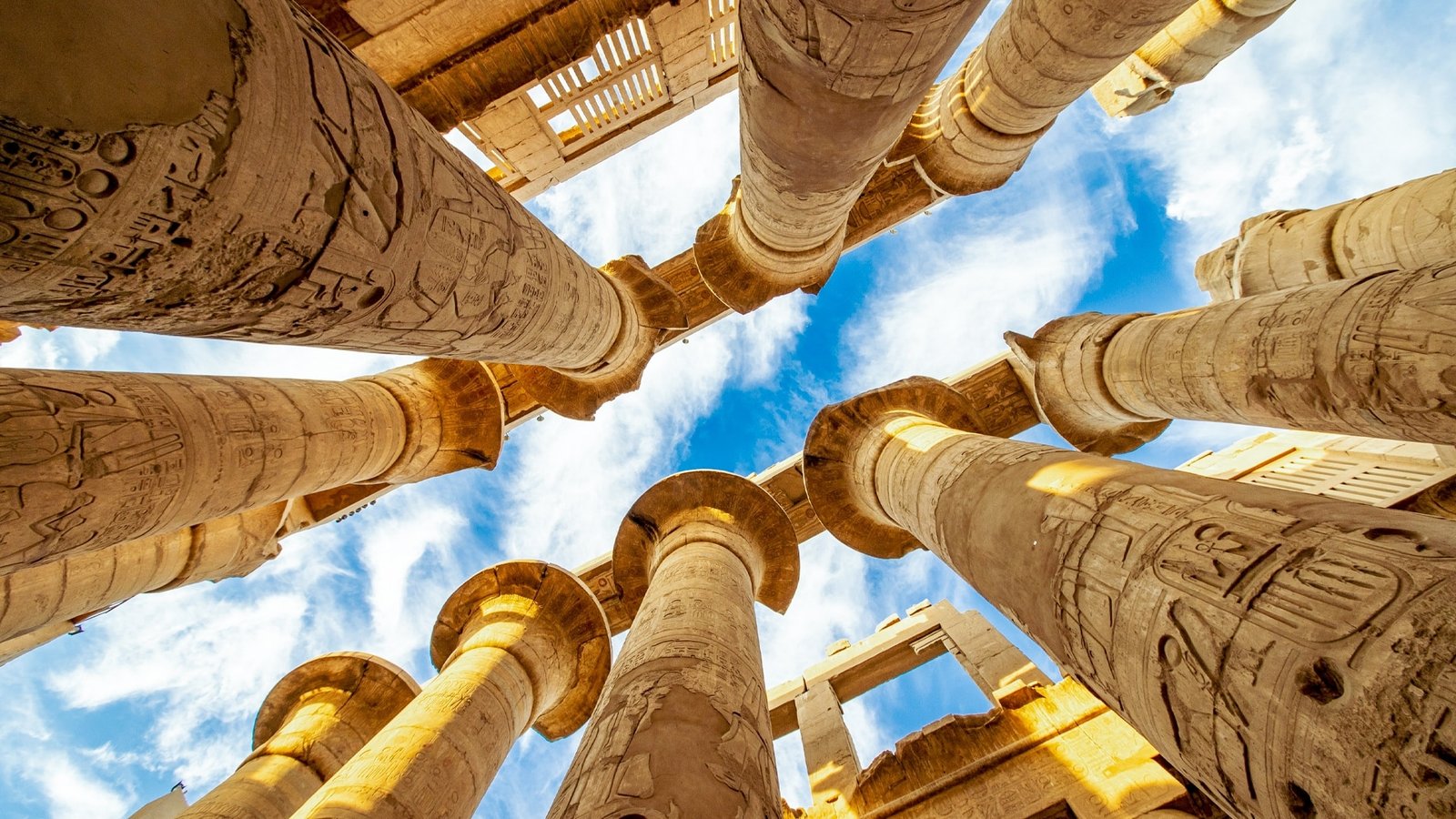 Carved sandstone columns and lintels of Karnak Templeโs hypostyle hall framing a blue sky with white clouds