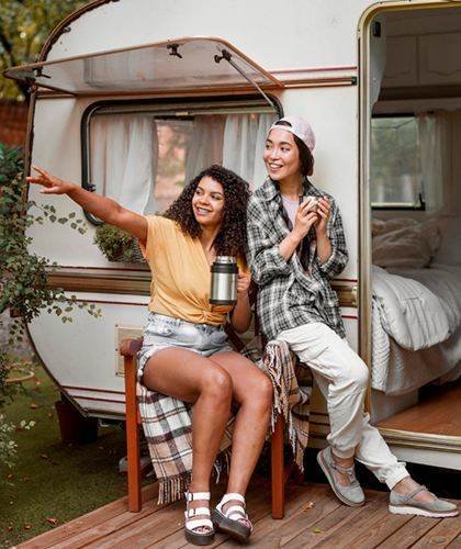 Two travelers seated beside an open camper trailer, with one pointing into the distance