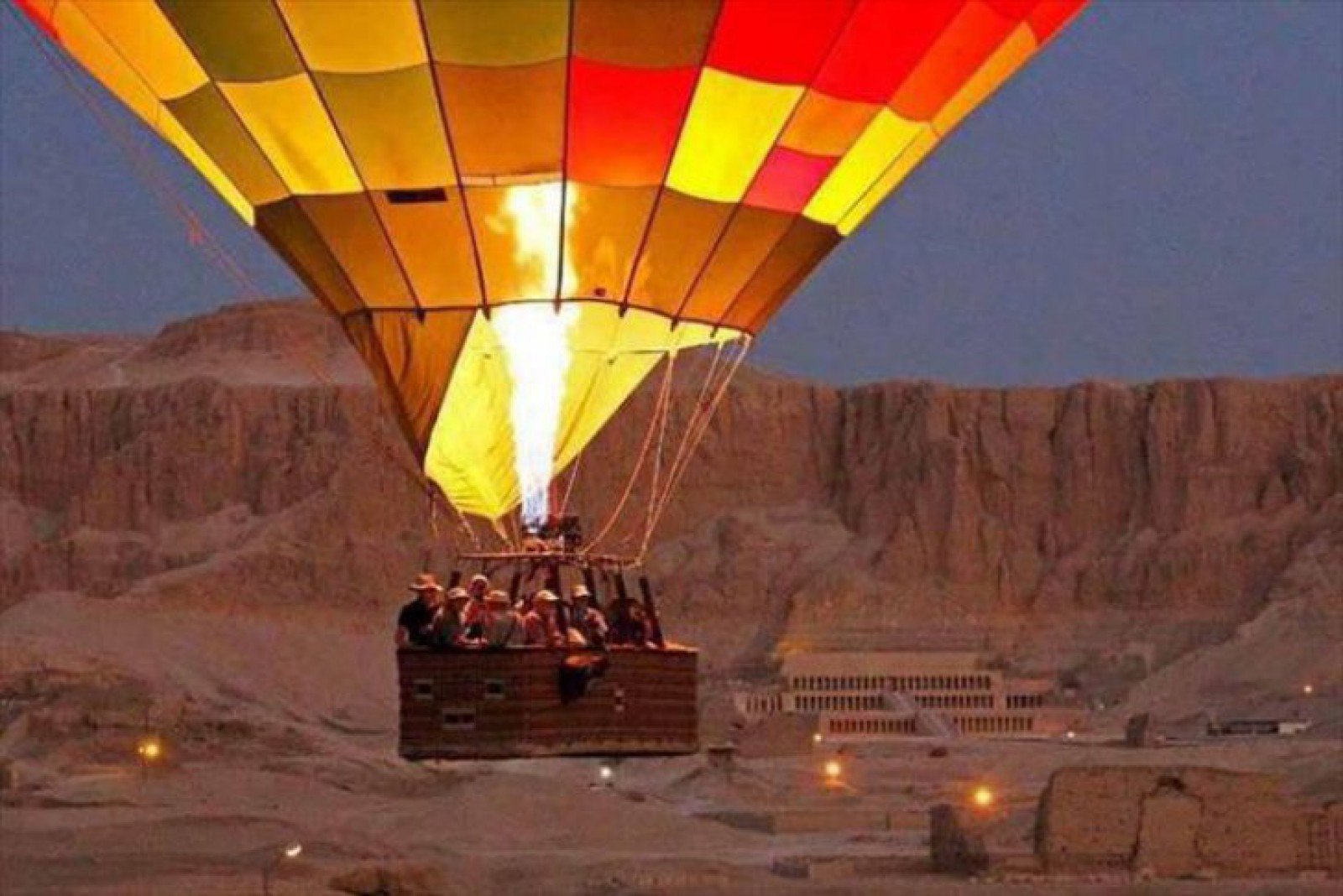 Passengers in a hot air balloon with a glowing burner above the west bank landscape near the Temple of Hatshepsut