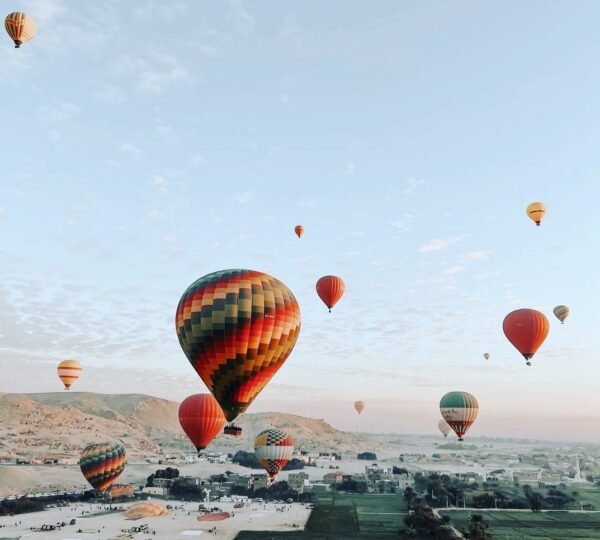 A large multicolored hot air balloon among smaller balloons above green fields, village buildings and desert hills in Luxor