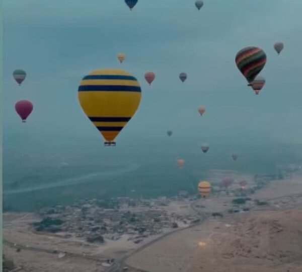 A yellow balloon with blue stripes and other balloons floating above villages, fields and desert roads near Luxor