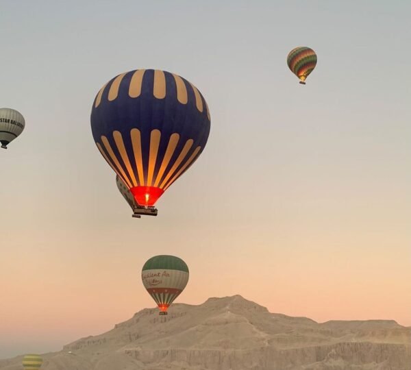 A blue and yellow hot air balloon glowing from its burner, with other balloons above Luxorโs desert hills
