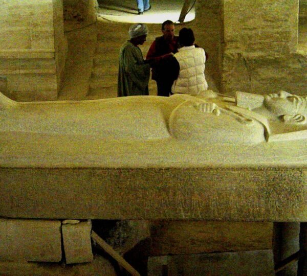 A stone sarcophagus with a carved reclining royal figure on its lid, with visitors standing behind