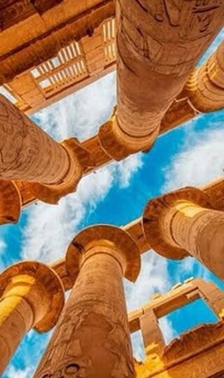 An upward view between carved sandstone columns toward a blue sky with white clouds at Karnak Temple