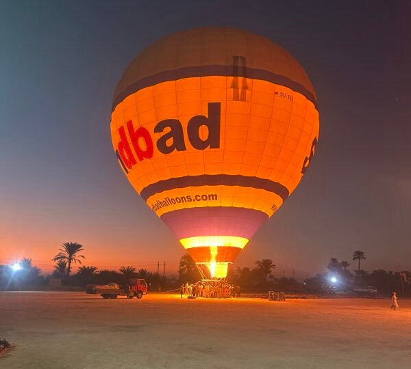 A hot air balloon glowing from its burner on the launch field before dawn in Luxor