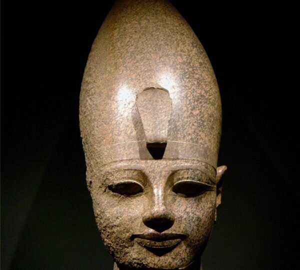 A carved granite head wearing a tall royal crown, illuminated against a dark museum background