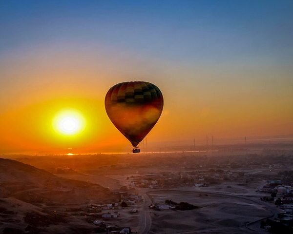 A colorful hot air balloon floating beside the rising sun above Luxorโs fields and desert hills