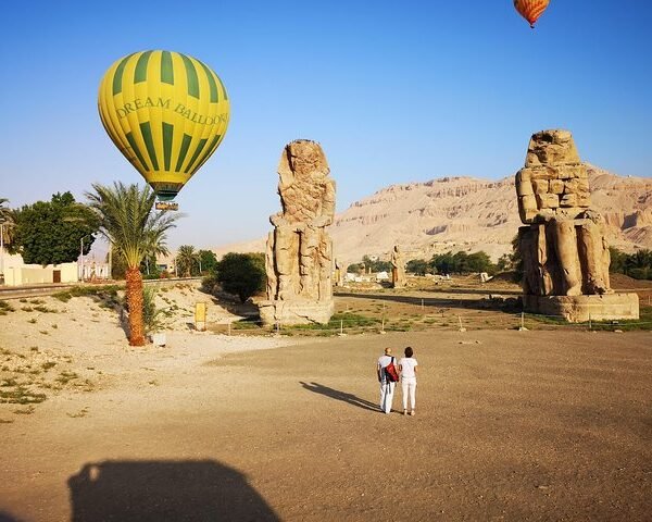 A yellow and green hot air balloon floating beside the Colossi of Memnon on Luxorโs west bank