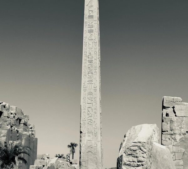 A monochrome photograph of a hieroglyph-covered obelisk rising above visitors and ancient stone ruins at Karnak