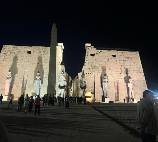 Visitors before the illuminated entrance pylons, statues and obelisk of Luxor Temple at night