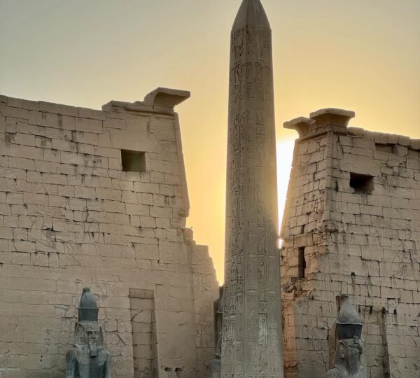 A hieroglyph-carved obelisk between Luxor Temple’s pylons and seated statues with the evening sun behind