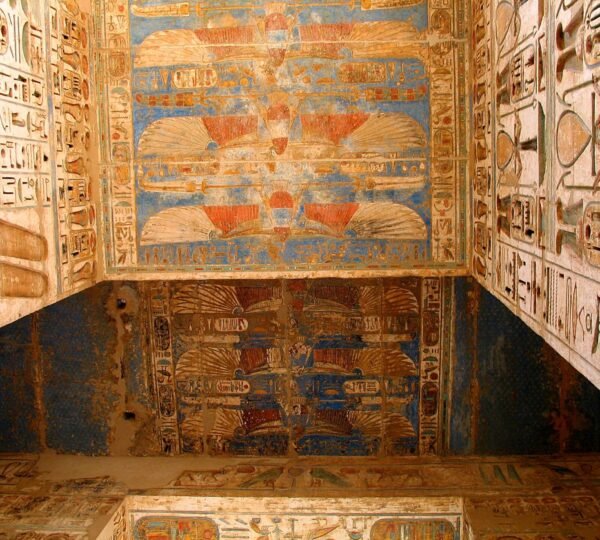 Red and gold winged figures on a blue painted ceiling bordered by carved hieroglyphs inside an Egyptian temple