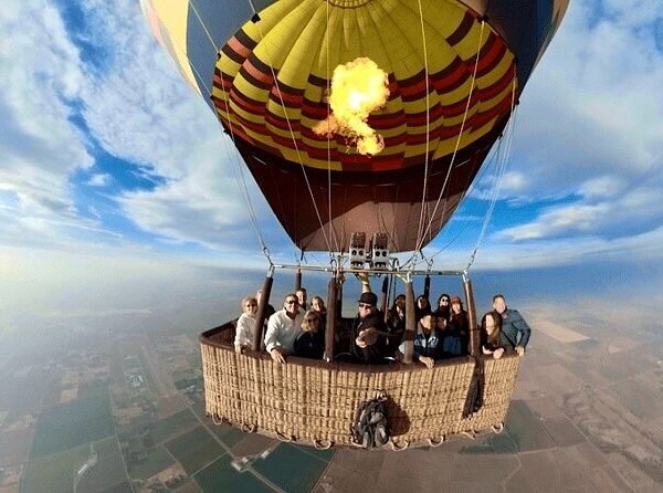 Passengers in a wicker balloon basket beneath a glowing burner, with fields and the Nile landscape below