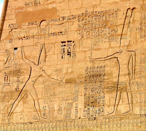 A large carved pharaoh with a raised arm facing another Egyptian figure, surrounded by hieroglyphs on a temple wall