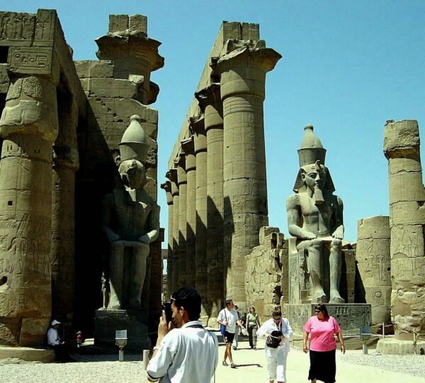 Visitors walking among seated royal statues and carved columns in a sunlit courtyard at Luxor Temple