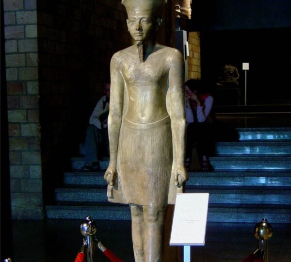 A crowned standing ancient Egyptian royal statue displayed behind a red barrier in a museum gallery