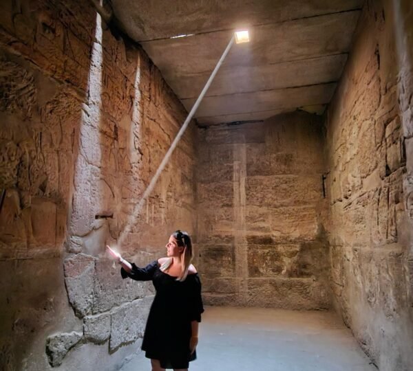 A visitor standing beneath a narrow sunbeam entering through the roof of a stone chamber covered in Egyptian reliefs