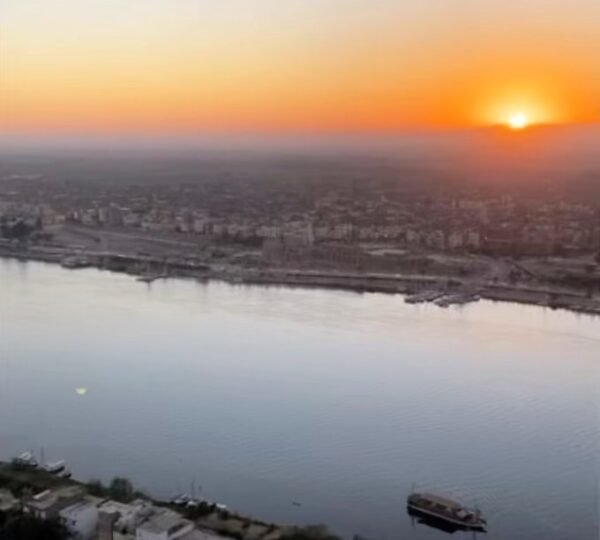 The rising sun above Luxorโs riverside buildings, with the Nile and a passenger boat below