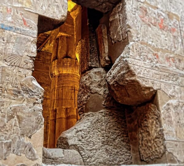 A warmly lit temple column seen through a rough opening in a wall covered with ancient Egyptian reliefs