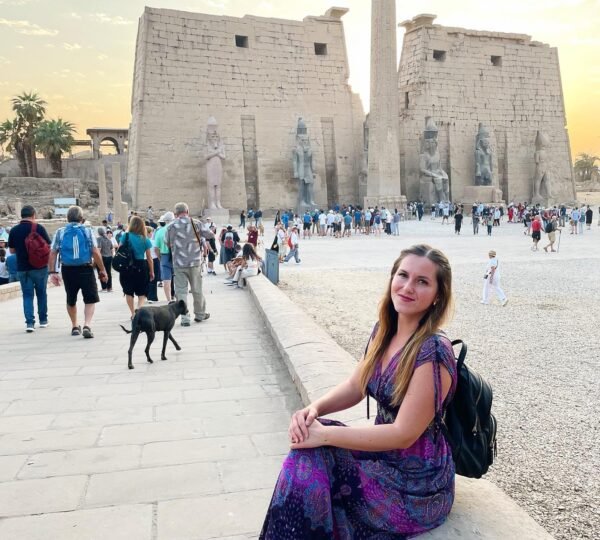 A traveler seated beside the stone path with Luxor Temple’s entrance pylons, obelisk and visitors behind