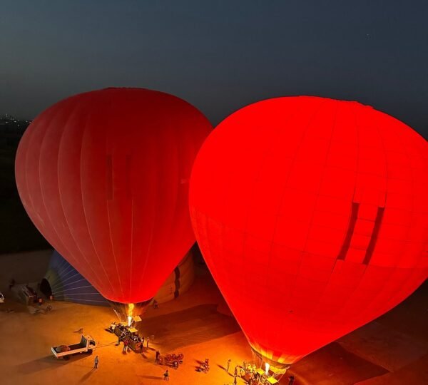 Two red hot air balloons glowing from their burners on the launch field, with ground crew and vehicles nearby
