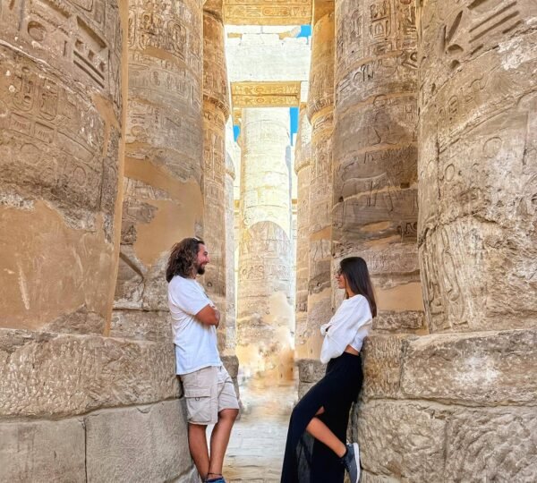 Two travelers standing between massive hieroglyph-covered columns in the hypostyle hall at Karnak Temple