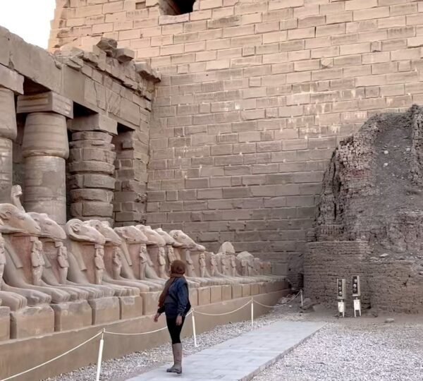 A visitor walking beside a row of ram-headed sphinxes beneath the entrance pylon of Karnak Temple
