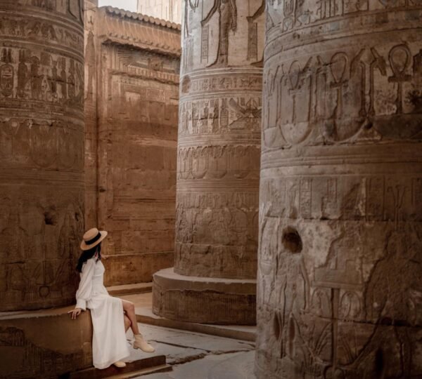 A visitor in a white dress seated beside large relief-covered columns inside Dendera Temple