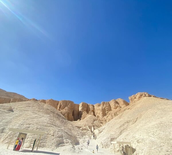 Visitors walking between tomb entrances and pale limestone hills in the Valley of the Kings beneath a clear blue sky