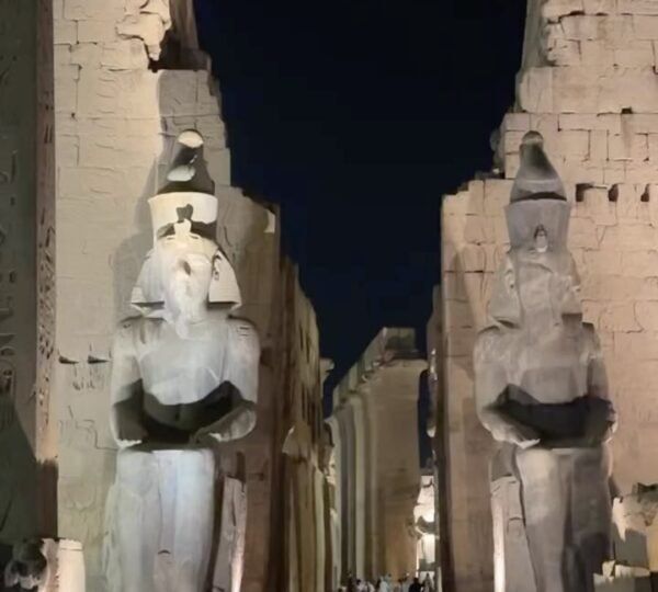 Visitors walking beneath two towering seated statues at the illuminated entrance of Luxor Temple at night