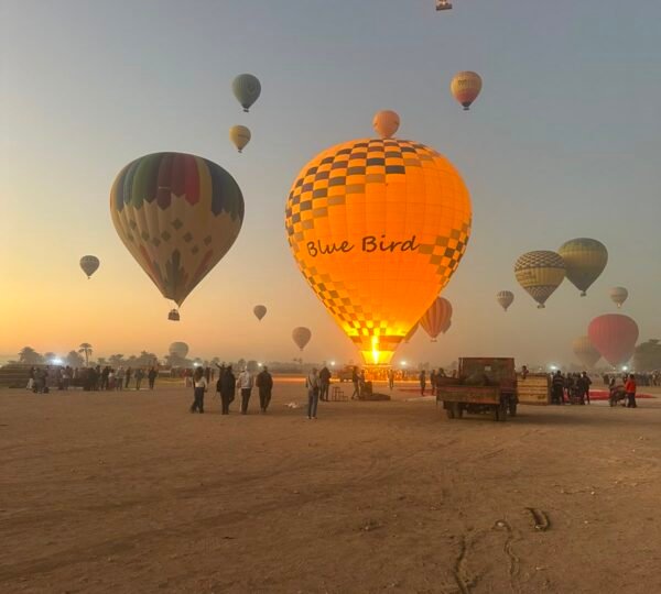 A yellow checkered balloon with a glowing burner among other balloons, passengers and crew on the Luxor launch field