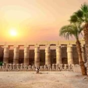 Ram-headed sphinxes and a stone colonnade at Karnak Temple beside palm trees beneath a warm evening sky