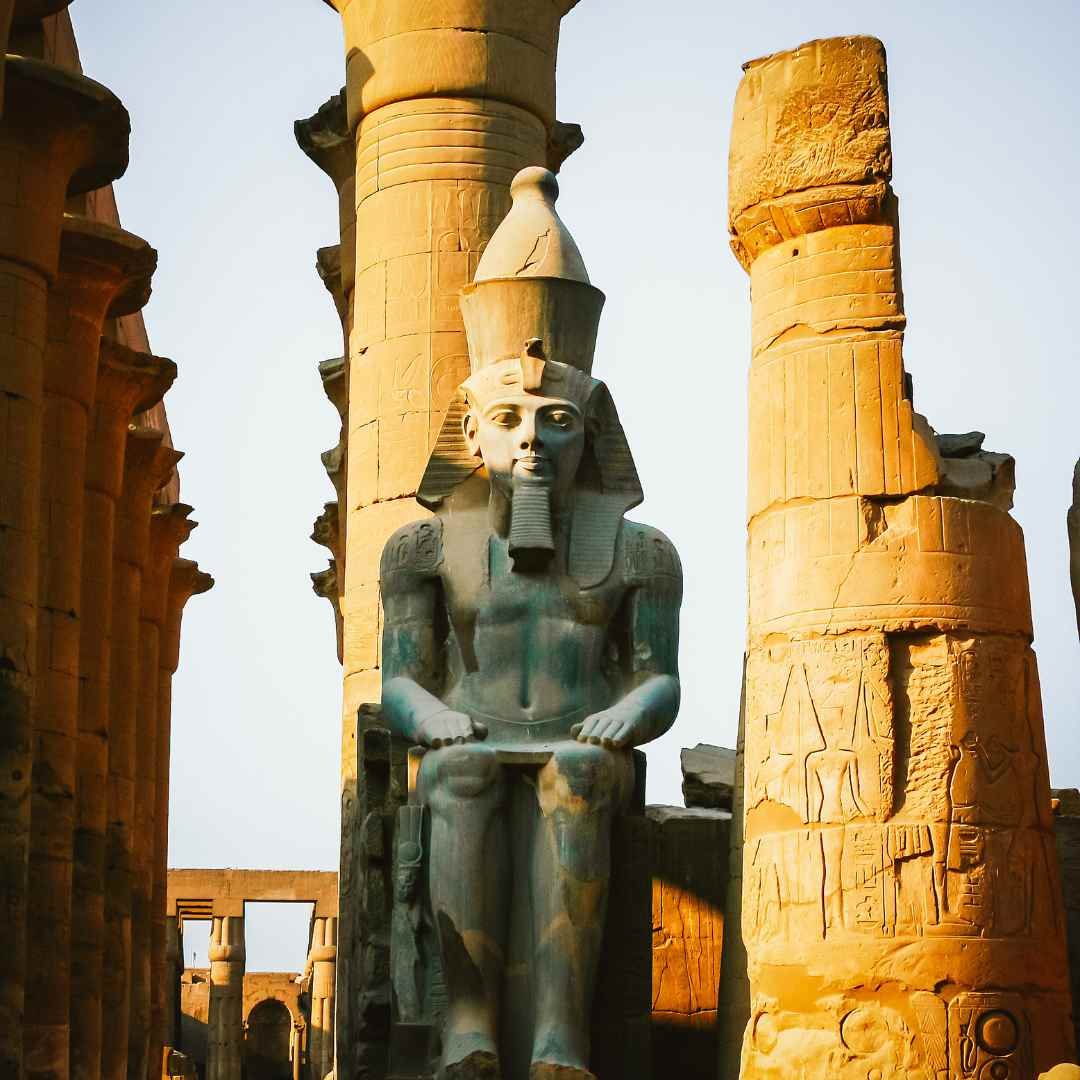 A seated royal statue wearing a crown and ceremonial beard among the sandstone columns of Luxor Temple