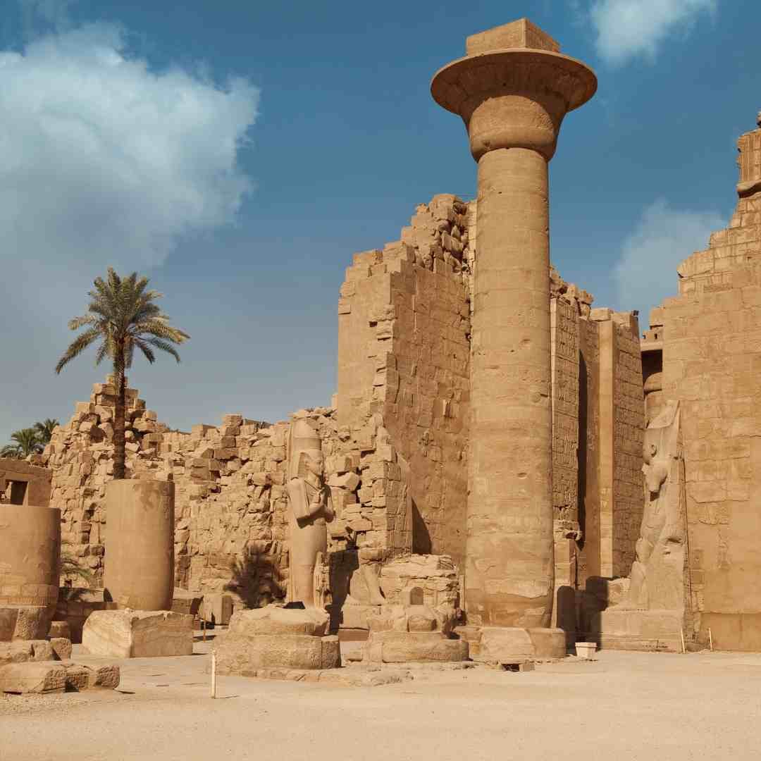 A tall papyrus-shaped column and standing statue among ancient stone ruins and palm trees at Karnak Temple