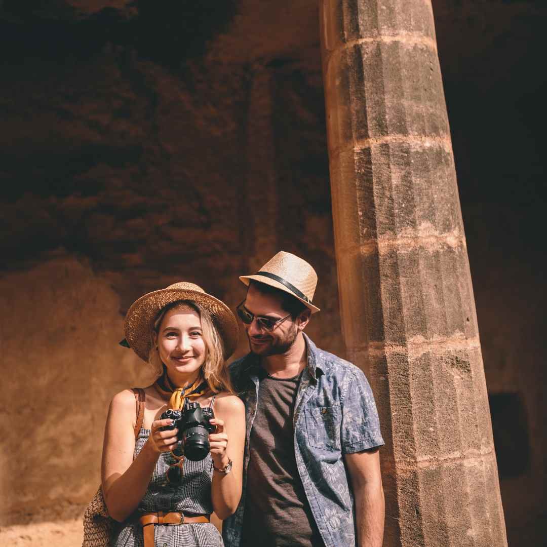 Two travelers wearing hats beside an ancient stone column, with one holding a camera