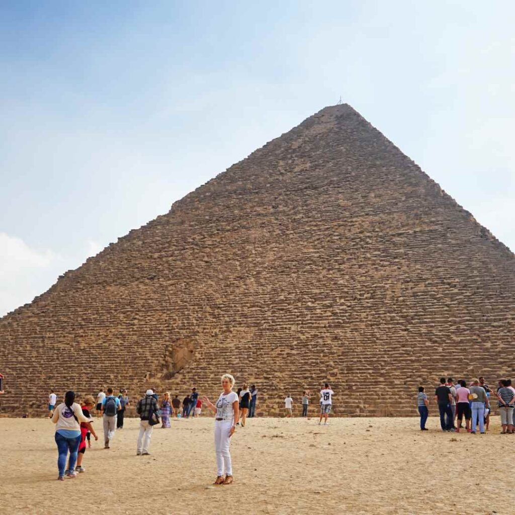 Visitors standing on the sandy ground beside the large stone-block face of a pyramid at Giza