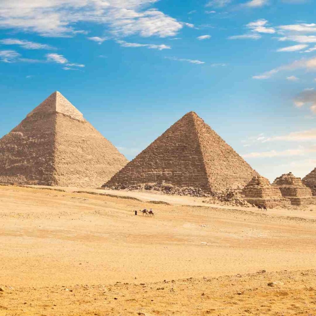 Two large pyramids and smaller pyramid ruins on the sandy Giza plateau beneath a blue sky with scattered clouds