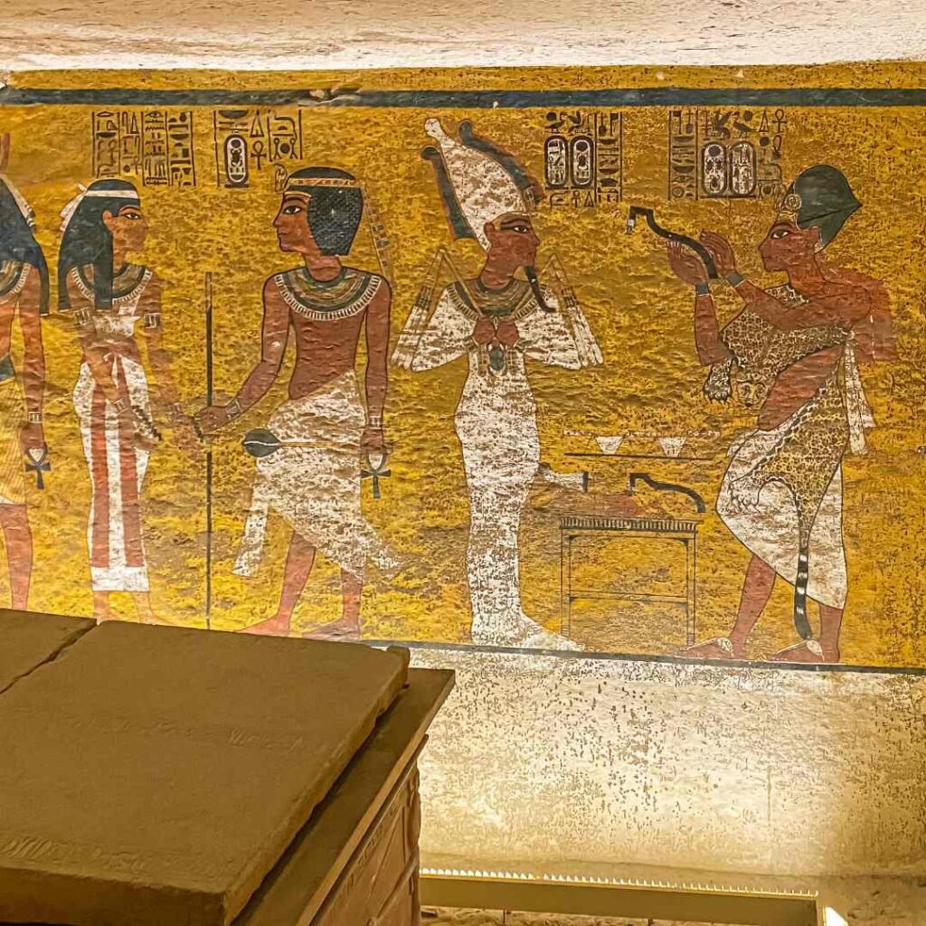 Painted Egyptian figures and hieroglyphs on a yellow tomb wall behind the edge of a stone sarcophagus