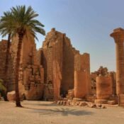 A palm tree beside ancient sandstone walls, broken columns and statues at Karnak Temple